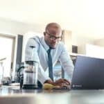 Geschäftsmann mit einem Laptop auf dem Küchentisch zu Hause. Mann arbeitet, während er frühstückt.